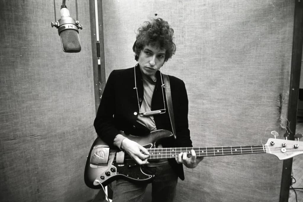 Bob Dylan in a recording studio, a harmonica rack around his neck, playing a bass guitar beneath a hanging studio microphone.