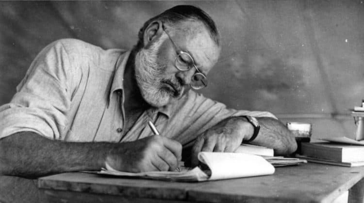 Ernest Hemingway seated at a wooden table, glasses low on his nose, writing in a notebook.