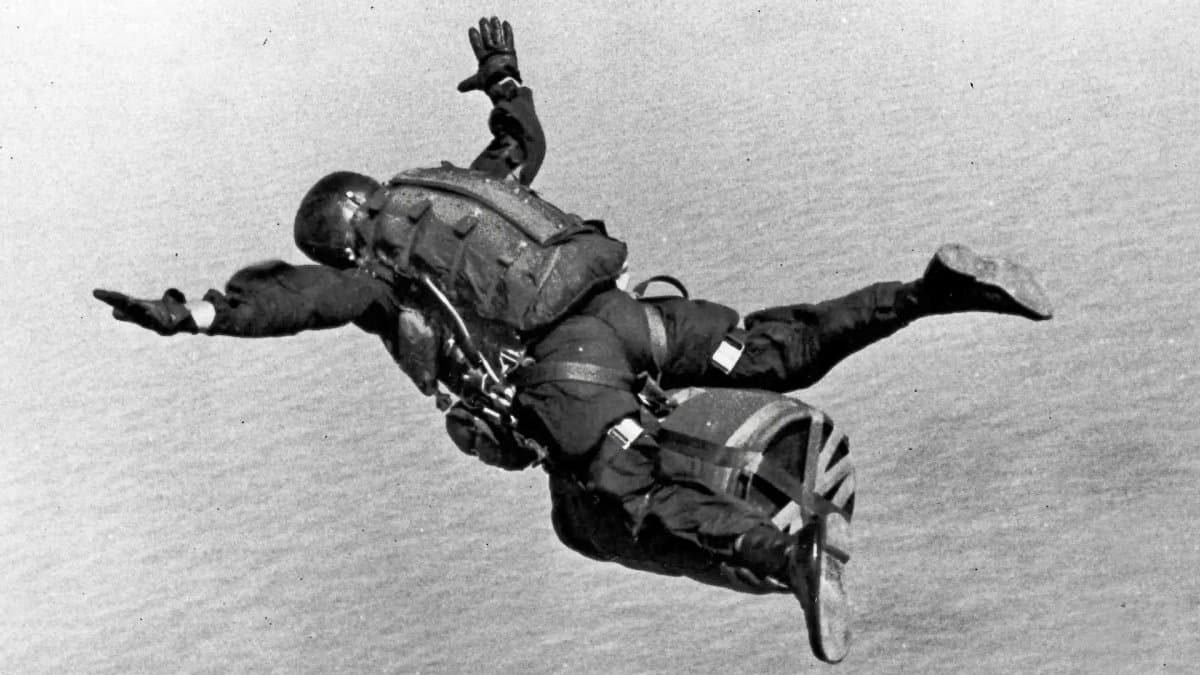 Black and white photograph of a military skydiver in freefall high above the water, arms spread wide, carrying a heavy pack.